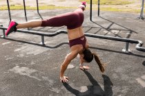 Vue de côté à angle élevé d'une femme sportive caucasienne avec de longs cheveux noirs faisant de l'exercice dans une salle de gym extérieure pendant la journée, faisant du handstand, les jambes tendues. — Photo de stock