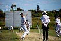 Vista trasera de un jugador de cricket masculino caucásico adolescente usando blancos, lanzando la pelota en el campo durante un partido de cricket, con un árbitro de pie detrás de él. - foto de stock