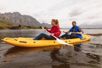Vista lateral de una pareja caucásica pasando un buen rato en un viaje a las montañas, kayak en un lago, con una cabaña en el fondo - foto de stock