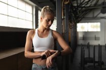 Vista frontal de una atlética mujer caucásica con ropa deportiva cruzando el entrenamiento en un gimnasio, de pie y mirando smartwatch, usando una aplicación de fitness - foto de stock