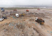 Disparo de drones de vehículos trabajando, limpiando y entregando basura apilada en un vertedero lleno de basura. Cuestión medioambiental mundial de la eliminación de residuos. - foto de stock