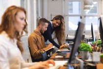 Homme d'affaires caucasien créatif travaillant dans un bureau moderne décontracté, assis à un bureau et souriant, prenant des notes avec des collègues féminines travaillant à côté de lui — Photo de stock