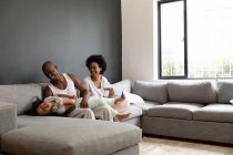 Vista frontal de una pareja afroamericana y su hija joven relajándose en la sala de estar juntos por la mañana, la pareja sentada en el sofá con la hija acostada en el regazo de su padre riendo - foto de stock