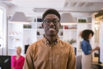 Porträt eines glücklichen afroamerikanischen Geschäftsmannes, der in einem modernen Büro arbeitet, in die Kamera blickt und lächelt, während seine Geschäftspartner im Hintergrund arbeiten — Stockfoto