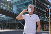 Homem caucasiano sênior nas ruas da cidade durante o dia, usando uma máscara facial contra coronavírus, vívido 19, usando um smartphone . — Fotografia de Stock