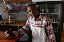 Afro-Américain travaillant dans un pub de microbrasserie, portant un tablier blanc, tenant une bouteille de bière et regardant droit dans une caméra. — Photo de stock