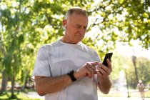 Vista frontal de um homem maduro caucasiano sênior trabalhando em um parque em um dia ensolarado, usando smartphone, se preparando para o treino — Fotografia de Stock