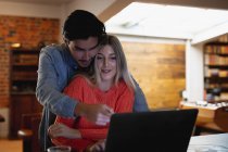 Vorderansicht einer jungen kaukasischen Frau und eines jungen Mannes mit gemischter Rasse, die die Zeit zu Hause genießen, in ihrem Wohnzimmer sitzen, lächeln und sich umarmen, während sie ihren Laptop benutzen.. — Stockfoto