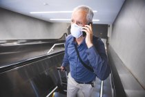 Hombre caucásico mayor, usando una máscara facial contra el coronavirus, covid 19, usando una escalera mecánica en una estación de metro, hablando en su teléfono inteligente, y tirando de una maleta. - foto de stock