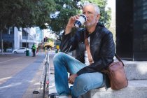 Senior Caucasian man, wearing casual clothes, out and about in the city streets during the day, sitting on stairs and holding a cup of takeaway coffee. — Stock Photo