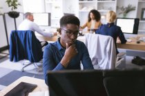 Um empresário afro-americano trabalhando em um escritório moderno, sentado em uma mesa e usando um computador, com seus colegas de negócios trabalhando em segundo plano — Fotografia de Stock