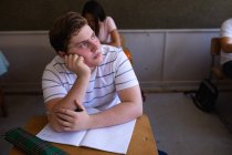 Високий кут перед очима Кавказького підлітка, який сидить за столом у шкільному класі, озираючись убік, думаючи, однокласники сидять за партами на задньому плані. — стокове фото
