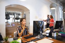Mixed race female business creative working in a casual modern office, sitting at a desk and using a computer, talking on a phone with colleagues working in the background — Stock Photo