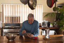 Vista frontal de un hombre caucásico mayor relajándose en casa, sentado en el mostrador en su cocina usando una tableta - foto de stock