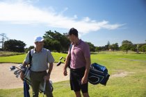 Vista frontal de dos hombres caucásicos en un campo de golf en un día soleado con cielo azul, caminando, llevando bolsas de golf, hablando - foto de stock