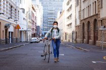 Vista frontal de una mujer de raza mixta con cabello oscuro en las calles de la ciudad durante el día, con una máscara facial contra la contaminación del aire y el coronavirus, caminando con su bicicleta con edificios en el fondo. - foto de stock