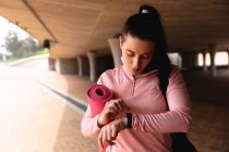 Vista frontale di una donna caucasica in forma sulla strada per l'allenamento fitness in una giornata nuvolosa, portando una borsa sportiva e un tappetino yoga, in piedi sotto un ponte, controllando il suo orologio — Foto stock