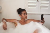 Смешанная расовая женщина проводит время дома самоизоляция и социальное дистанцирование в карантинной изоляции во время эпидемии коронавируса ковид 19, лежа в ванной расслабляясь, используя свой смартфон в ванной комнате. — стоковое фото