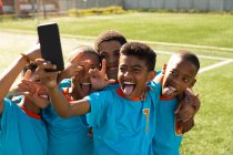 Front view close up of a multi-ethnic group of boy soccer players wearing their team strip, standing on a playing field taking a selfie with a smartphone, and posing with arms around each other, smiling and making faces — Stock Photo