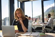Eine kaukasische Geschäftsfrau, die in einem modernen Büro arbeitet, am Schreibtisch sitzt und einen Laptop benutzt, während ihre Kollegen im Hintergrund arbeiten — Stockfoto