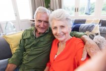 Portrait d'un heureux couple caucasien retraité à la maison dans leur salon, assis sur un canapé, regardant à la fois la caméra et souriant, la femme prenant un selfie, couple isolant pendant la covid19 coronavirus pandémie — Photo de stock