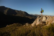 Seitenansicht des kaukasischen Mannes, der die Zeit in der Natur genießt, Reißverschlussausrüstung trägt, Wandern, der Mann steht auf einem Felsen an einem sonnigen Tag in den Bergen — Stockfoto