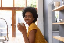 Vista laterale di una donna afro-americana a casa, seduta in cucina con in mano una tazza di caffè con la testa rivolta verso la macchina fotografica, distogliendo lo sguardo sorridente — Foto stock