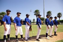 Vue de côté d'un groupe multi-ethnique de joueurs de baseball masculins, se préparant avant un match, debout dans une rangée, se préparant à chanter un hymne national — Photo de stock