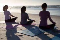 Vista trasera de un grupo multiétnico de amigas disfrutando haciendo ejercicio en una playa en un día soleado, practicando yoga sentadas en posición de yoga, meditando en posición de loto, frente al mar. - foto de stock