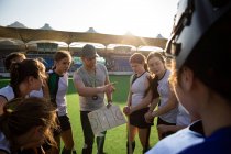 Seitenansicht einer Gruppe kaukasischer Hockeyspielerinnen, die sich vor einem Spiel auf einem Hockeyfeld um ihren kaukasischen männlichen Trainer schmiegen, der ihnen an einem sonnigen Tag einen Spielplan gibt und auf etwas zeigt. — Stockfoto