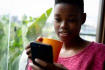 Nahaufnahme einer afroamerikanischen Frau, die an einem sonnigen Tag in ihrem Wohnzimmer vor einem Fenster sitzt, ein Smartphone benutzt und einen Becher in der Hand hält — Stockfoto