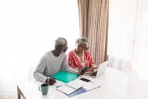 Una coppia afroamericana anziana che trascorre del tempo a casa insieme, distanziamento sociale e isolamento in quarantena durante l'epidemia di coronavirus covid 19, seduta a un tavolo, usando un computer portatile, l'uomo che tiene uno smartphone — Foto stock