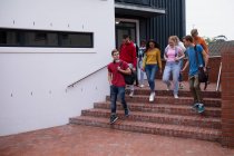 Front view of a multi-ethnic group of teenage male and female high school pupils with schoolbags hanging out, talking and walking down steps in their school grounds — Stock Photo