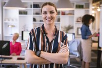 Portrait of a happy Caucasian businesswoman working in a modern office, looking at camera and smiling, standing with arms crossed, with her business colleagues working in the background — Stock Photo