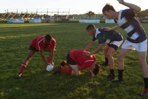Vista lateral de duas equipes masculinas multiétnicas adolescentes de jogadores de rugby usando suas tiras de equipe, em ação durante uma partida em campo de jogo, ajudando um jogador ferido a cair no chão — Fotografia de Stock