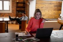 Vorderansicht einer jungen kaukasischen Frau, die im Wohnzimmer sitzt und während der Arbeit ihren Laptop benutzt, während ihr Partner im Hintergrund sitzt. — Stockfoto