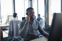 Un hombre de negocios afroamericano que trabaja en una oficina moderna, sentado en un escritorio, usando una computadora, usando auriculares y hablando, con sus colegas trabajando en el fondo - foto de stock