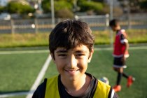 Portrait close up of a confident mixed race boy soccer player wearing a team strip, standing on a playing field in the sun, looking to camera and smiling, with a teammate in the background — Stock Photo