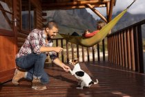 Vista laterale di una coppia caucasica che fa un buon viaggio in montagna, un uomo si inginocchia su un balcone in una cabina, giocando con i cuccioli, mentre una donna è sdraiata su un'amaca con un cucciolo sulla pancia. — Foto stock