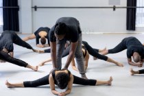 Front view of a mixed race fit male modern dancer wearing black outfit, supporting a female dancer while stretching up during a dance class in a bright studio, with other dancers exercising in the background. — Stock Photo