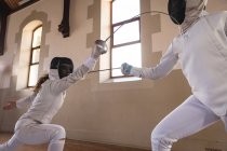 Caucasian and African American sportswomen wearing protective fencing outfits during a fencing training session, taking aim at each other with their epees. Fencers training at gym. — Stock Photo