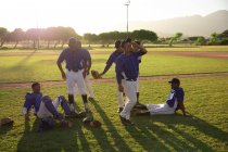 Vue latérale d'un groupe multiethnique de joueurs de baseball masculins, s'entraînant avant un match, se reposant, s'asseyant, interagissant, par une journée ensoleillée — Photo de stock