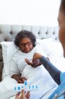 Старша змішана жінка проводить час вдома, лежачи в ліжку, відвідуючи змішану расу жінку-медсестру, медсестру, яка дає свої ліки — стокове фото