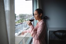 Vista lateral de una mujer de raza mixta con el pelo corto relajándose en casa, de pie en la sala de estar mirando por la ventana, bebiendo una copa de vino tinto - foto de stock