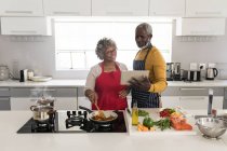 Ein älteres afroamerikanisches Paar, das während der Coronavirus-Epidemie 19 Zeit zu Hause verbringt, soziale Distanzierung und Selbstisolierung in Quarantäne, steht in der Küche und bereitet Essen zu, der Mann hält eine Tablette — Stockfoto