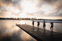 Передній вигляд веслувальної групи з чотирьох кавказьких жінок, які несуть човен на плечах, стоячи на пристані в річці на світанку. — стокове фото