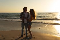 Kaukasisches Paar, das bei Sonnenuntergang am Strand steht, Händchen hält, einander umarmt und anschaut — Stockfoto