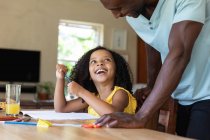 Chica afroamericana con una blusa amarilla, distanciamiento social en casa durante el cierre de cuarentena, sentada junto a una mesa y dibujando con su padre. - foto de stock