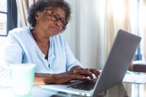 Старша змішана жінка насолоджується своїм часом вдома, соціальною дистанцією та самоізоляцією в карантині, сидячи за столом, використовуючи ноутбук — стокове фото