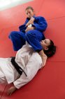 Vista frontale ad alto angolo di due adolescenti judoka caucasici e misti di razza femminile che indossano judogi blu e bianchi, che praticano il judo durante uno sparring in una palestra. — Foto stock